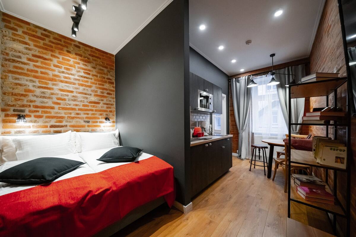 Una habitación con pared de ladrillo, cama con colcha roja, kitchenette con microondas y zona de comedor con mesa y sillas.