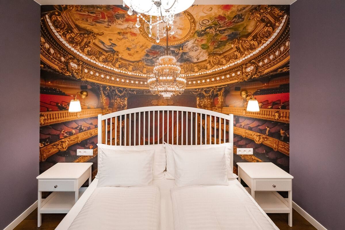 Un dormitorio con una cama grande, dos mesitas de noche y un mural que representa el techo de un teatro.
