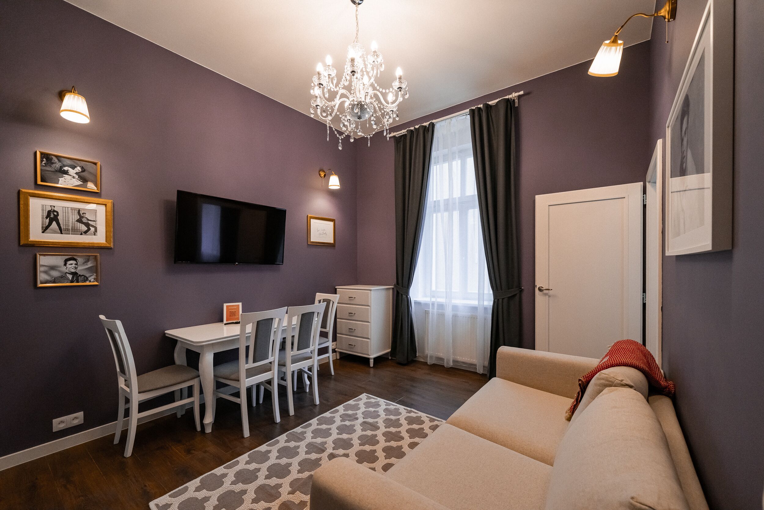 A modern living room with a sofa, a dining table with chairs, a TV mounted on the wall, and a chandelier.