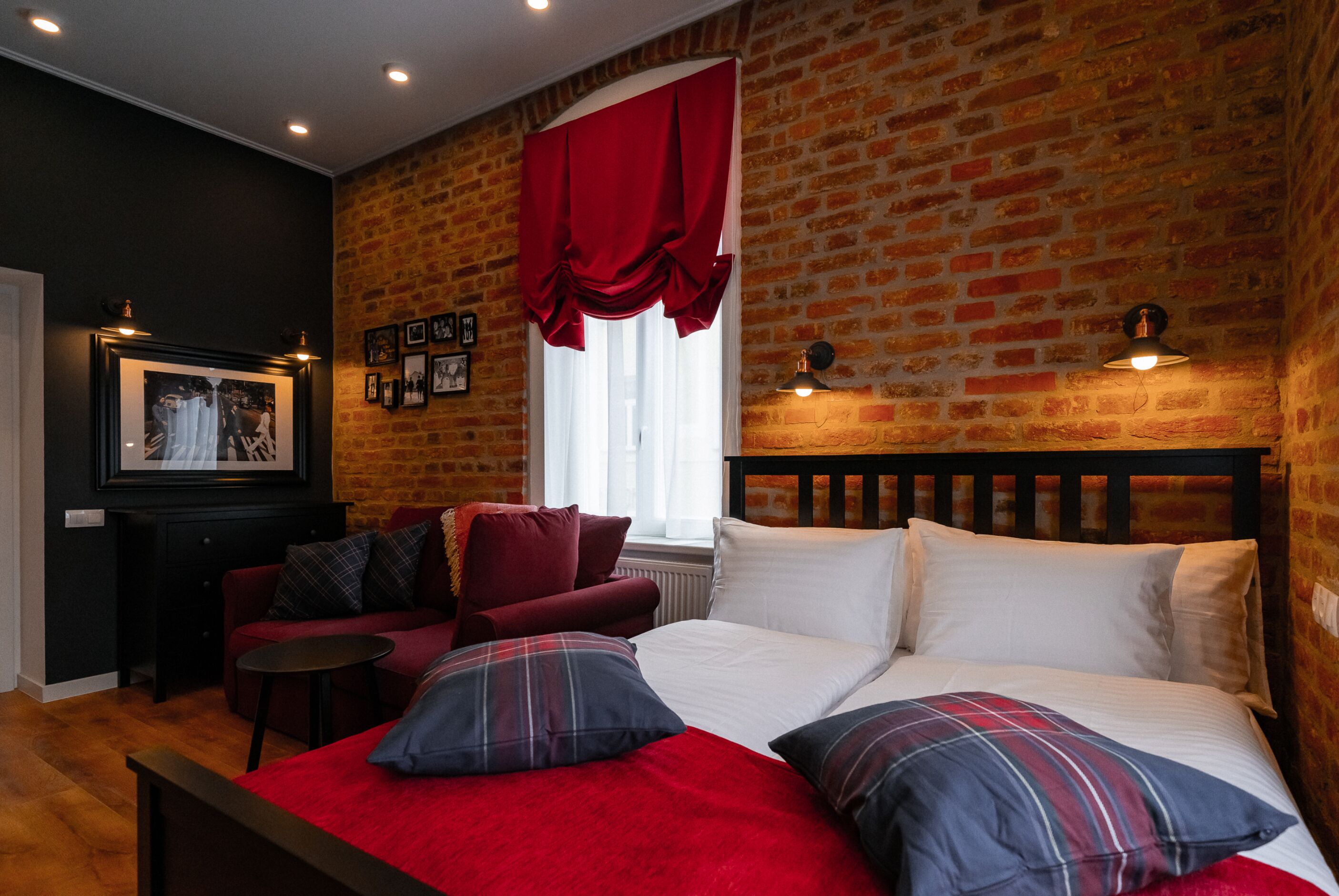 A hotel room with a brick wall, a bed with white and plaid bedding, a red sofa, and a small table.