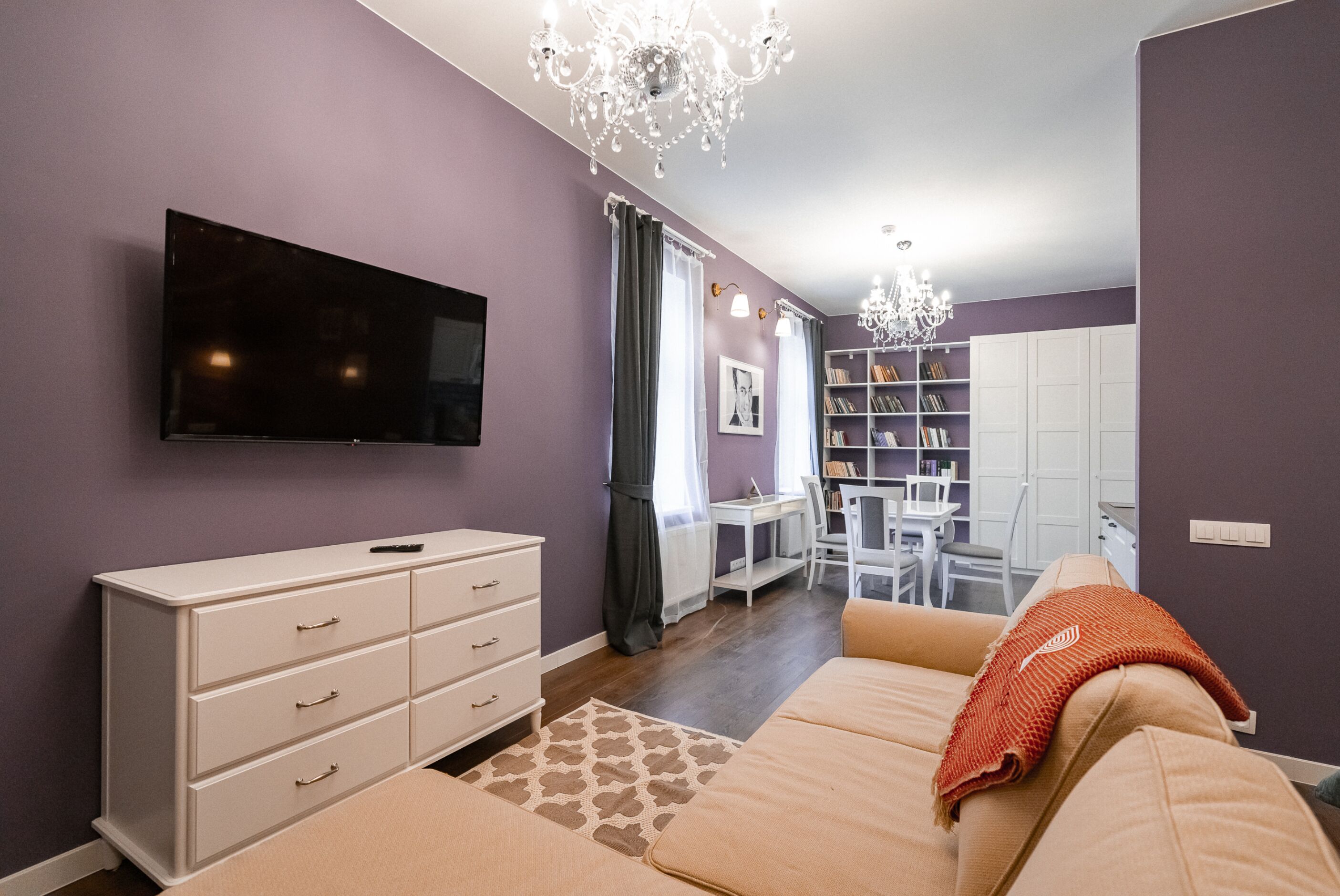 A modern living room with a flat-screen TV, a sofa, a white dresser, and a dining area with a table and chairs.
