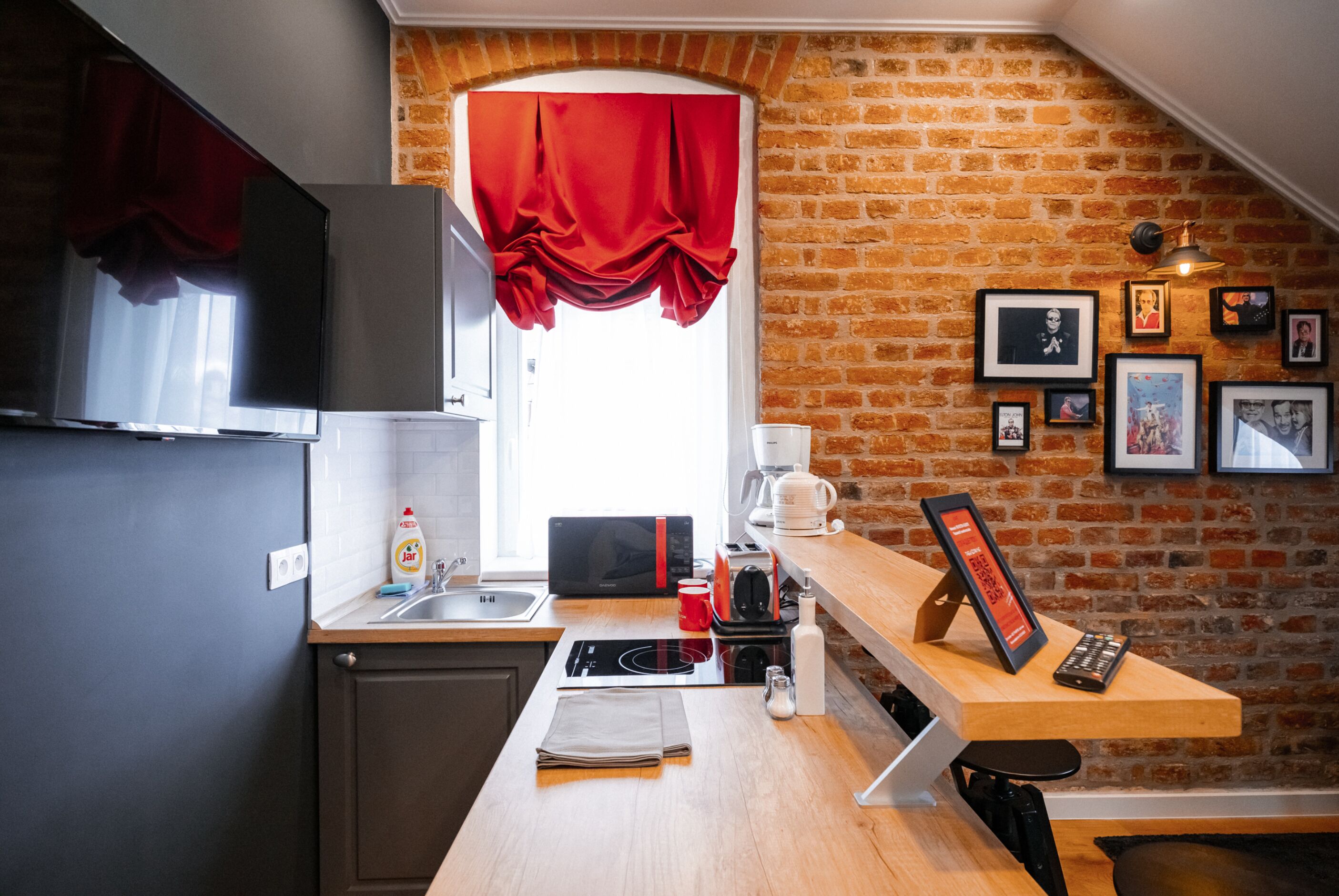 A kitchen with a brick wall, a window with red curtains, a sink, a microwave, a coffee maker, and framed pictures on the wall.