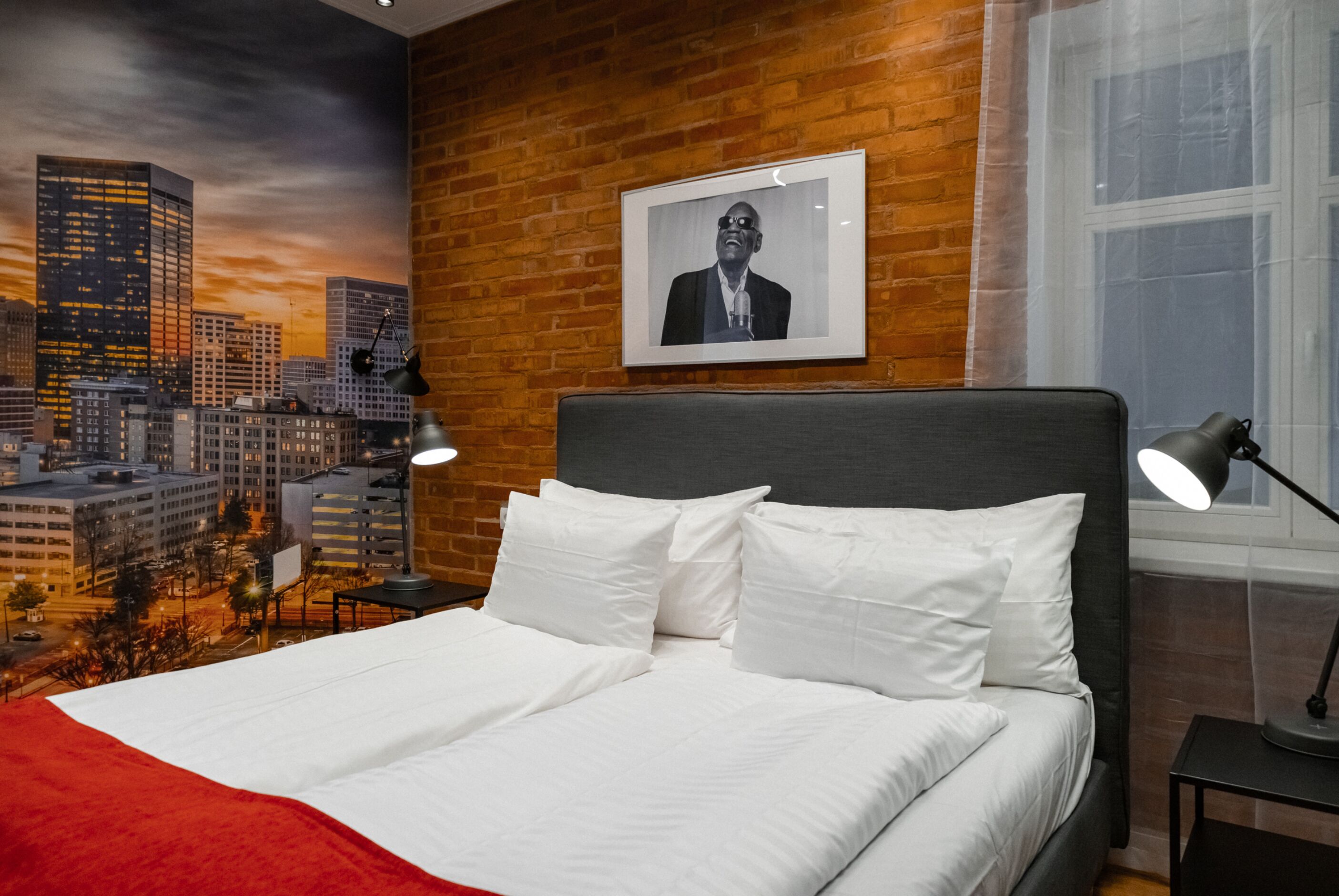 Ein Hotelzimmer mit Stadtbild-Wandgemälde, einem Bett mit weißen Bettwäsche, einem schwarzen Kopfteil und einem gerahmten Bild an der Wand.