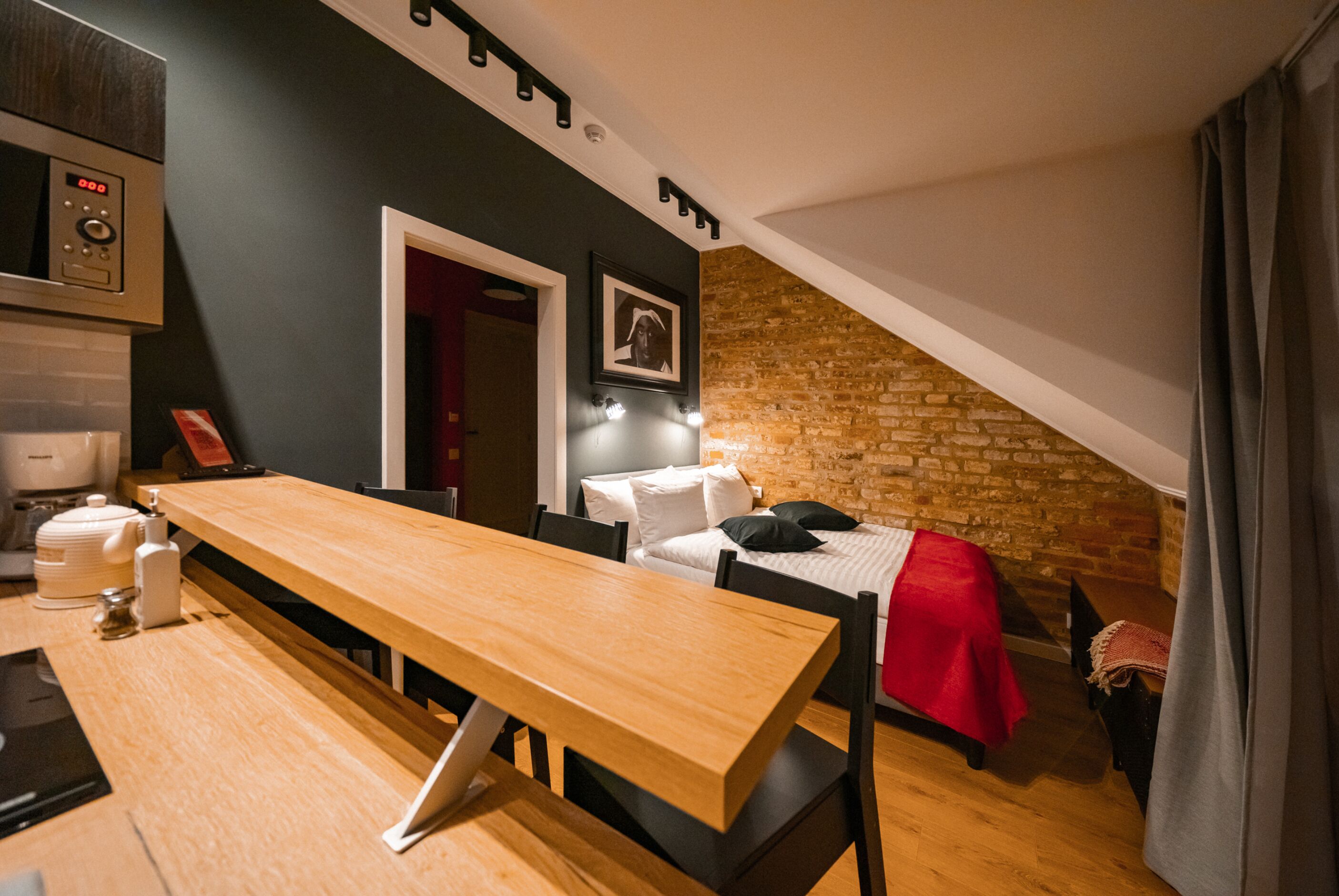 A compact room with a wooden dining table, a bed with a red blanket, a brick wall, and a kitchen area with a microwave.