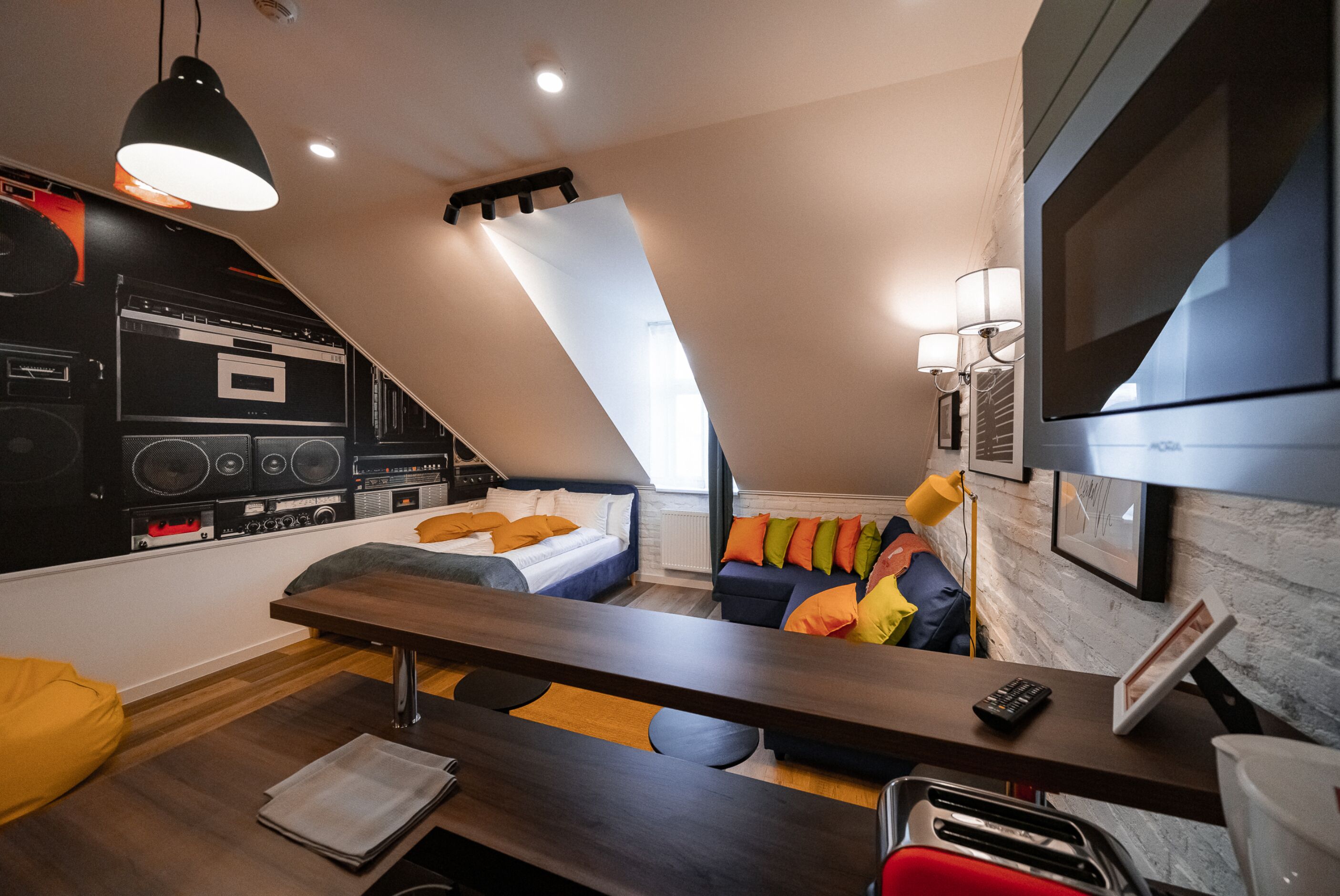 A modern loft bedroom with a sloped ceiling, a kitchenette, a large bed, a dining table, and a TV mounted on the wall.