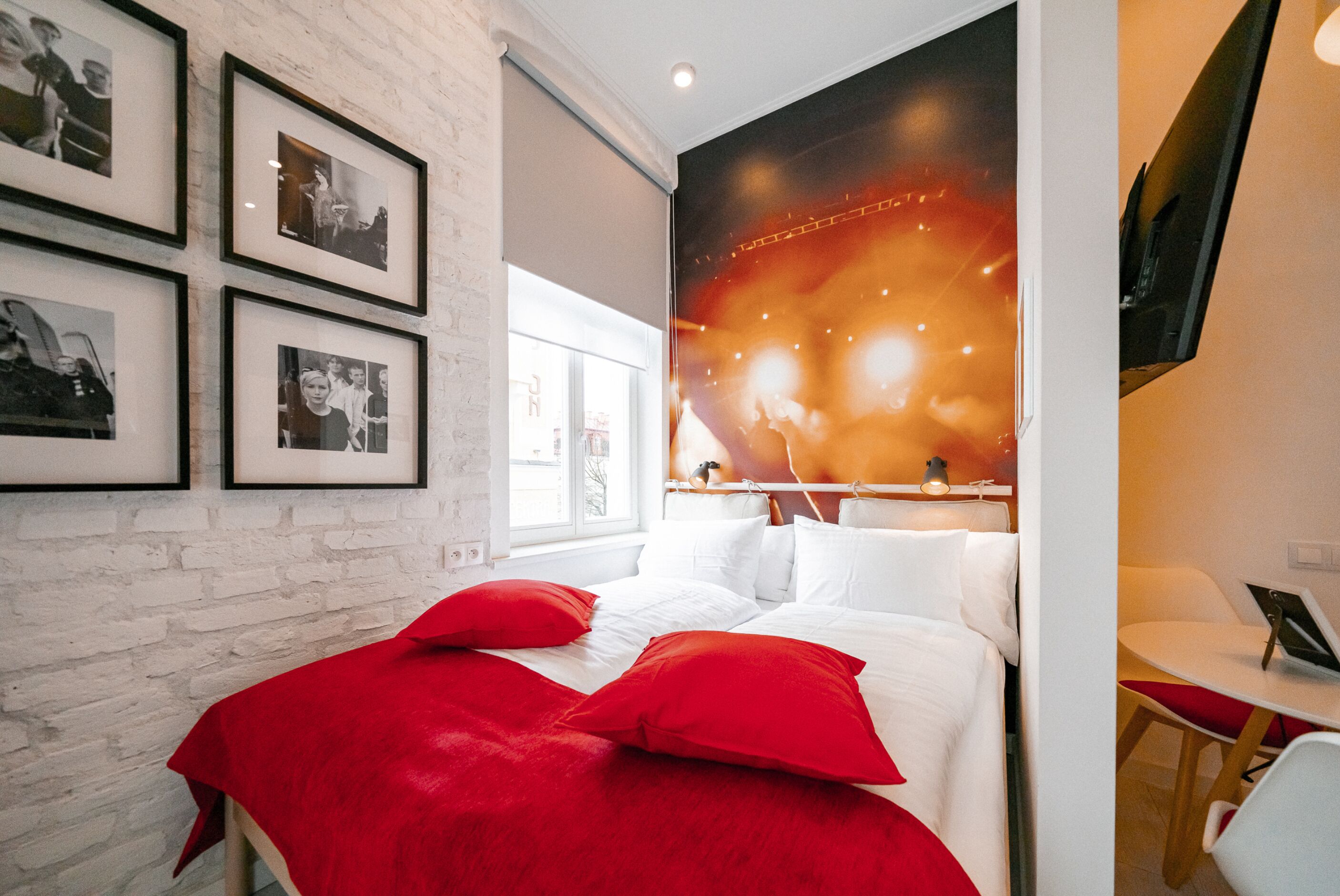 Une chambre d’hôtel moderne avec un mur en briques blanches, un grand lit avec une literie rouge, un téléviseur à écran plat et des photographies encadrées accrochées au mur.