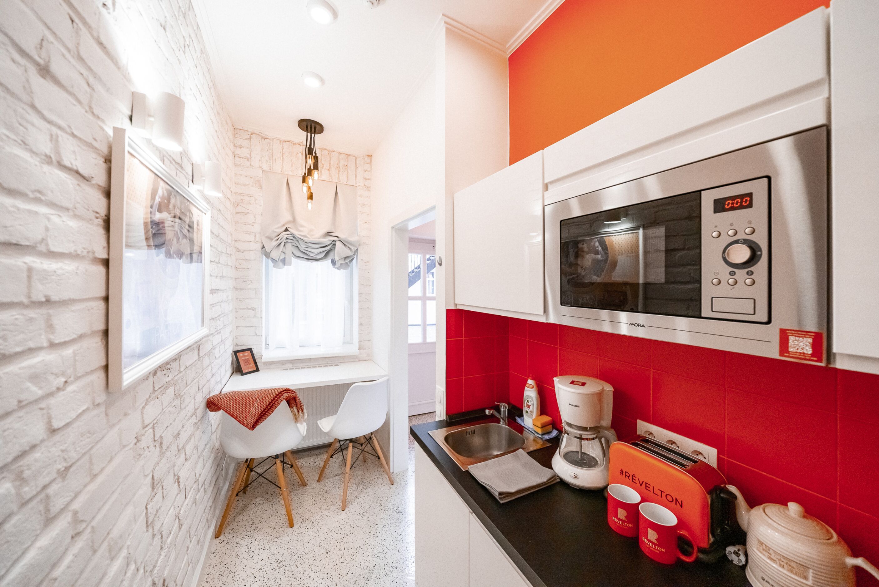 A kitchen with a microwave, toaster, and coffee maker. There is a window with a view of the outside, a white chair, and a red wall.