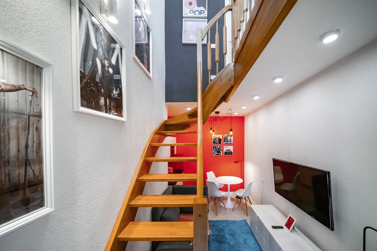 Un interior moderno con una escalera de madera, una pared de acento roja, una mesa redonda con sillas y un televisor montado.