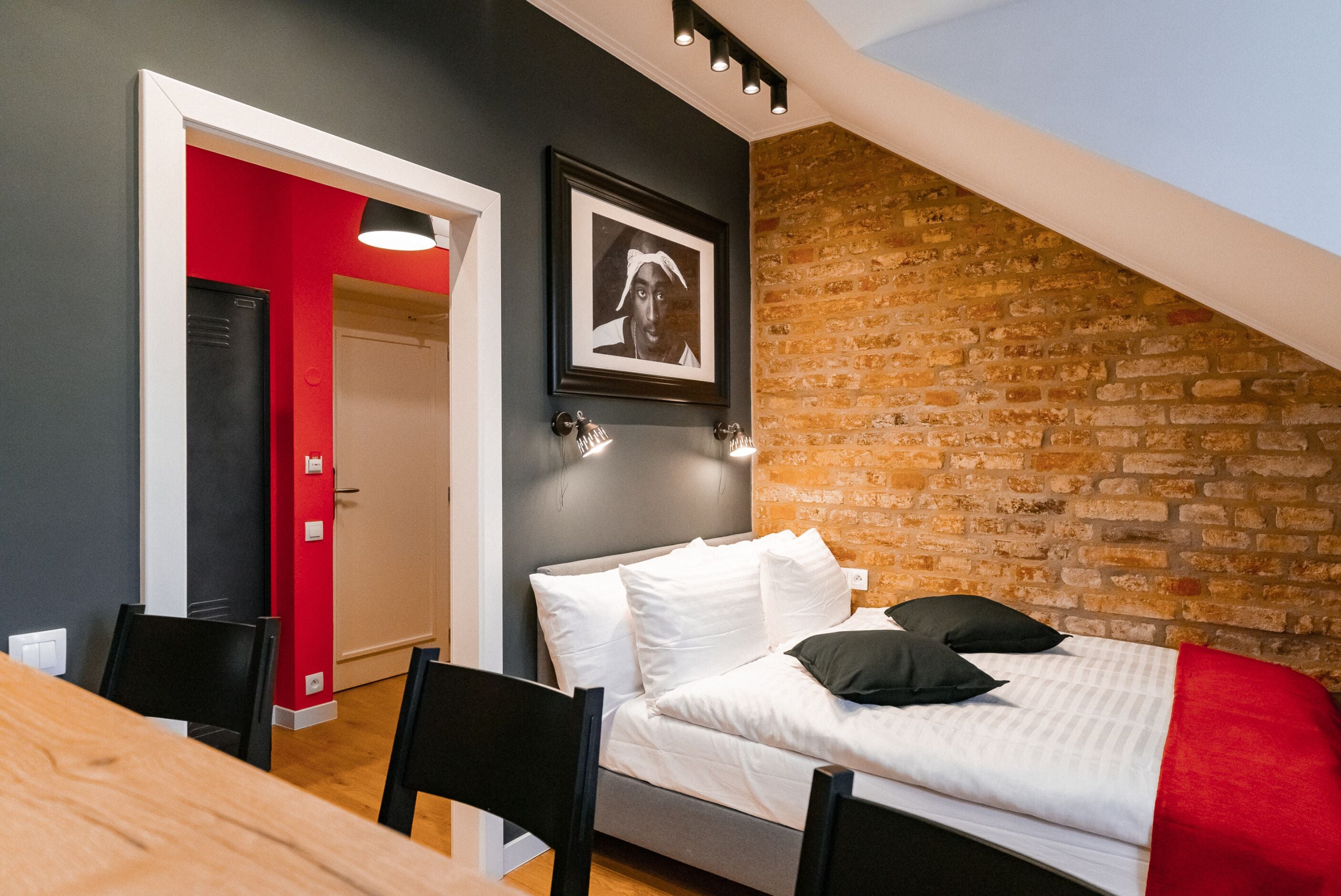 Ein Schlafzimmer mit einem Bett, einem Esstisch, Stühlen, einer roten Wand, einer Backsteinwand und einem gerahmten Bild an der Wand.
