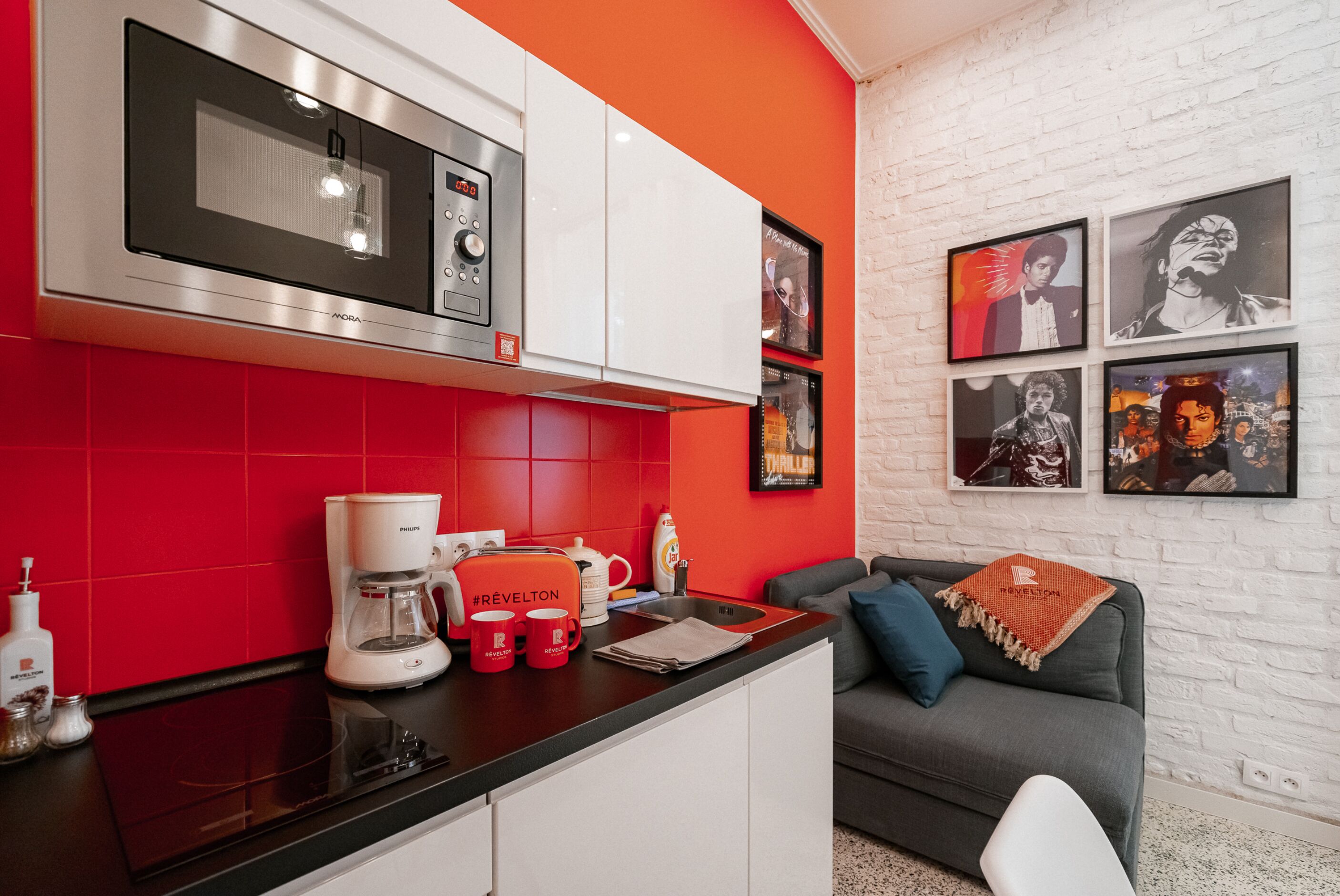 A modern kitchen with a red backsplash, white cabinets, and a black countertop. There is a coffee maker, mugs, and a kettle on the counter.