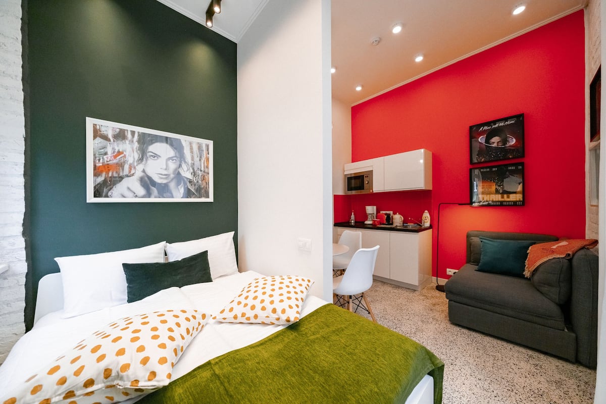 Un dormitorio moderno con una cama, una manta verde y un cojín con lunares. Hay una kitchenette con microondas y una pared roja.