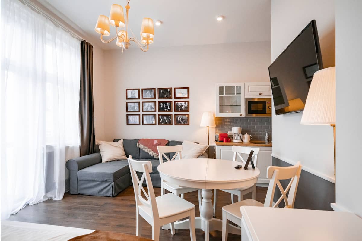 Una sala de estar moderna con un sofá gris, sillas blancas, una mesa redonda y una zona de cocina con televisor, microondas y cafetera.