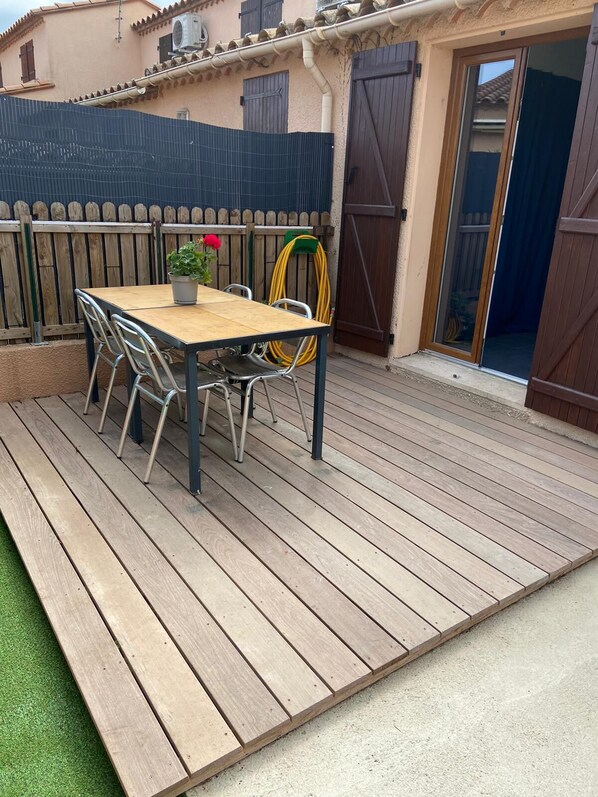 Une terrasse en bois avec une table et des chaises, un pot de fleurs et un tuyau d’arrosage.