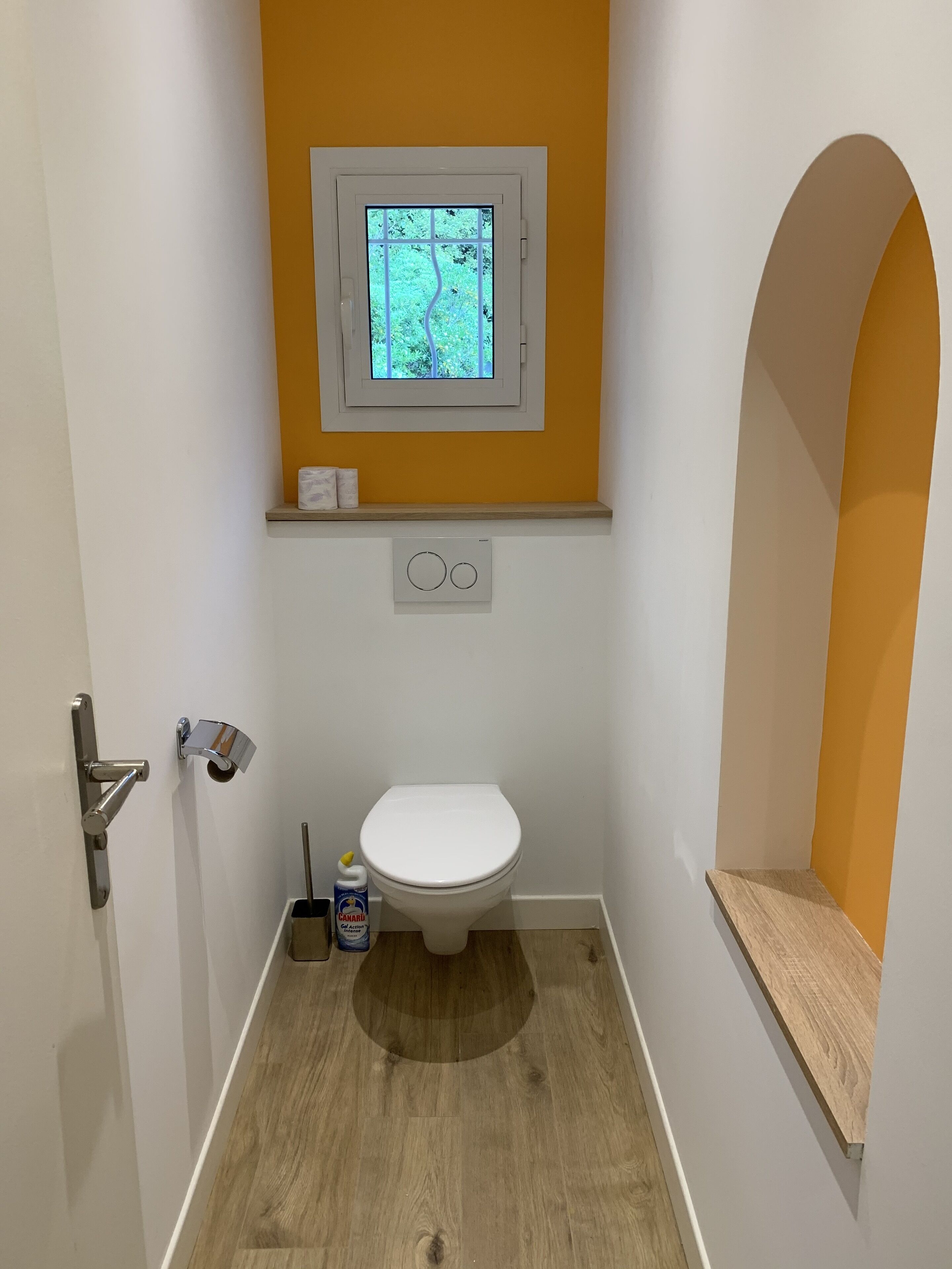 A small bathroom with a toilet, a window, and a wooden shelf.