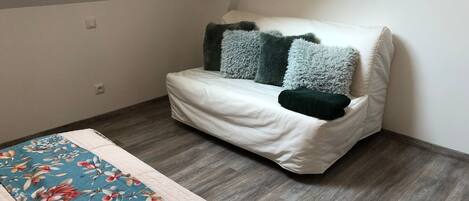 A small, cozy room with a white sofa against a sloped ceiling, a floral-patterned bedspread, and a wooden floor.