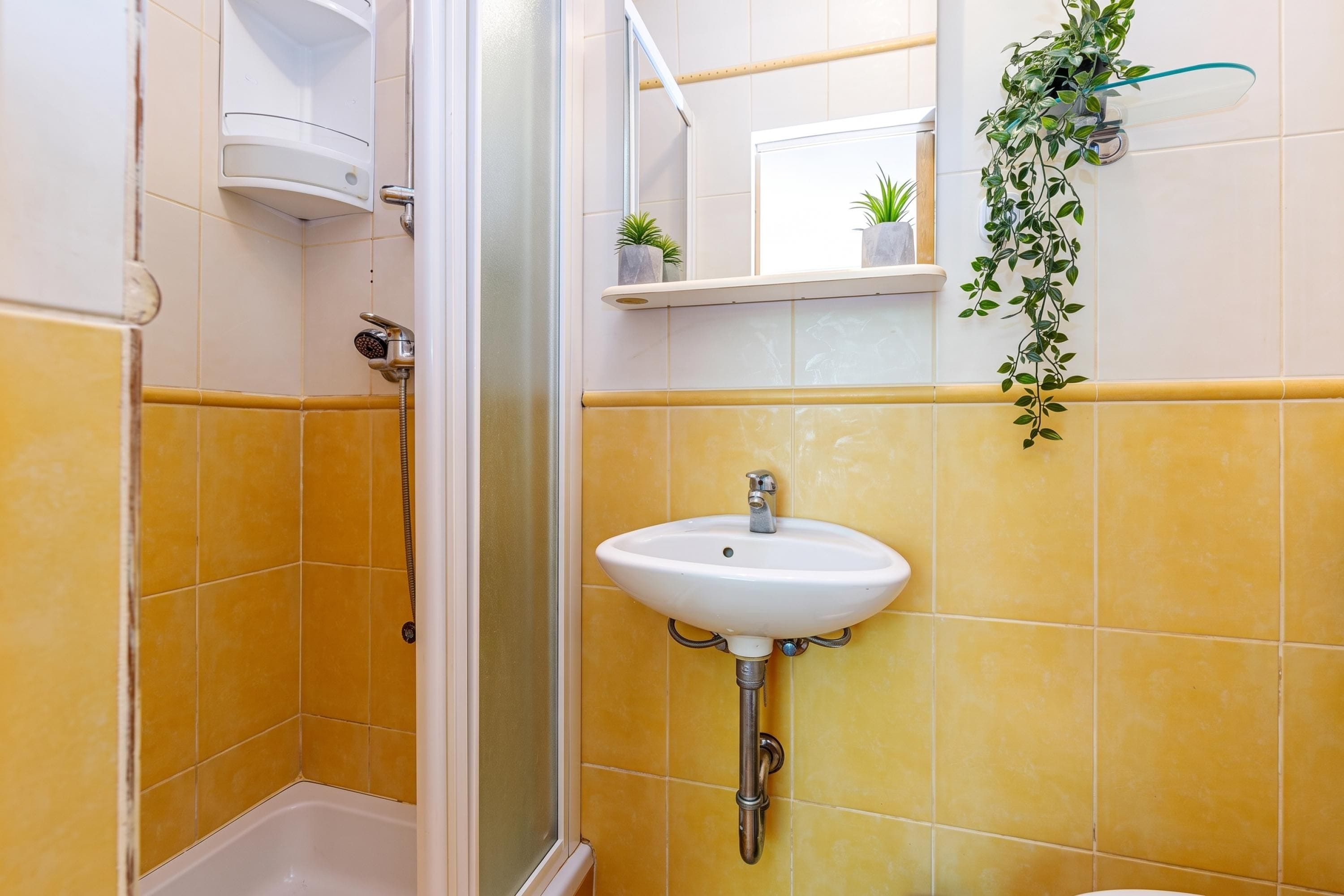 Une salle de bain avec des carreaux jaunes, un lavabo blanc, une cabine de douche en verre et une petite plante en pot posée sur une étagère.