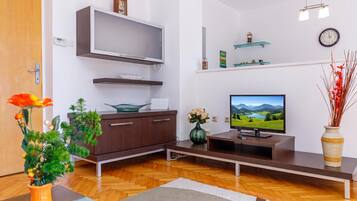A modern living room with a flat-screen TV, wooden flooring, a sofa, a coffee table, a vase with flowers, and a wall-mounted shelf with decorative items.