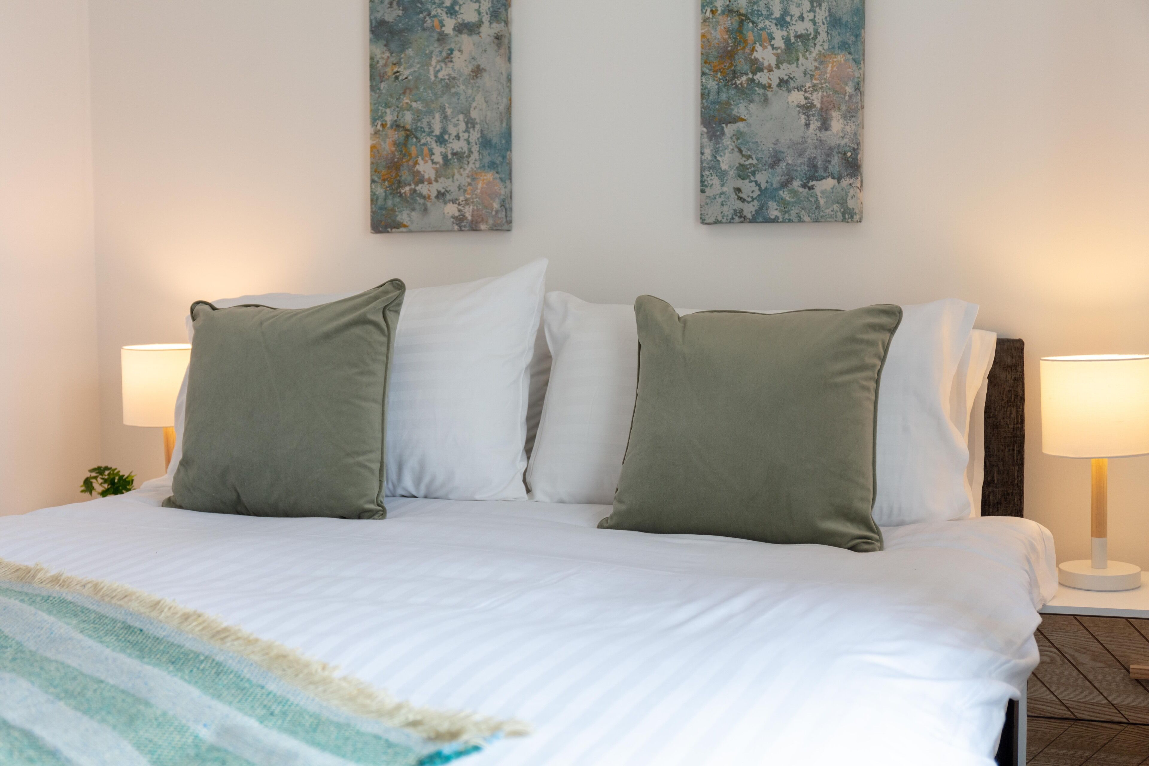 A neatly made bed with white linens and two green pillows, flanked by two bedside lamps on either side.