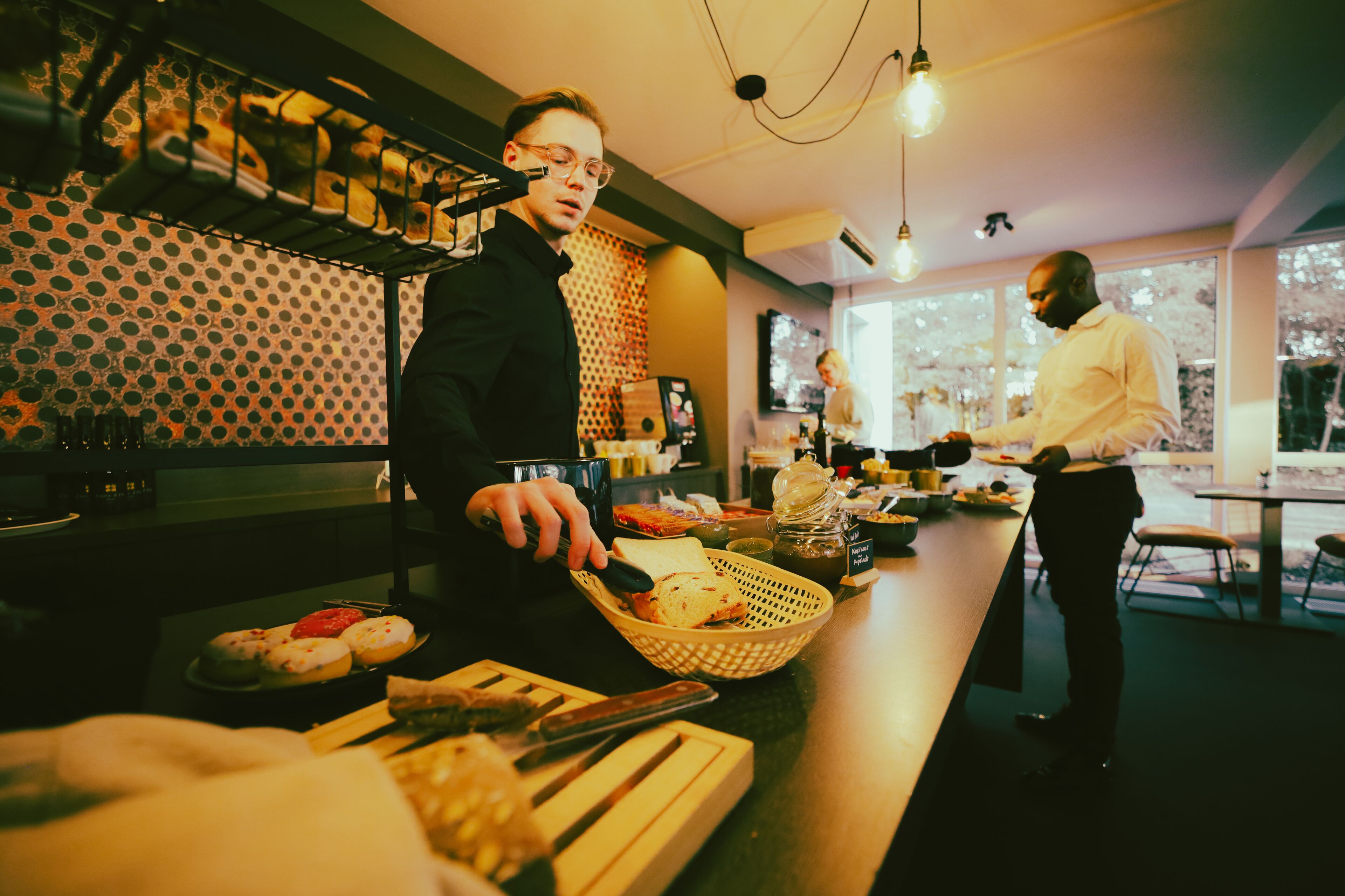 A person serving food at a counter with a patterned wall and large windows in the background.