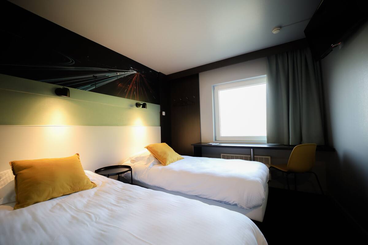 Hotellihuone, jossa on kaksi sänkyä, ikkuna, jossa on verhot, tuoli ja seinä, jossa on valaisin.