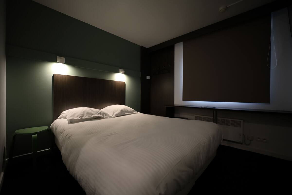 Hotellihuone, jossa on sänky, pieni pöytä, seinälle asennettu televisio ja seinälle asennettu valaisin.