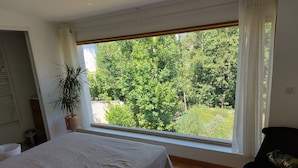 A bedroom with a large window, a bed, a chair, and a potted plant.