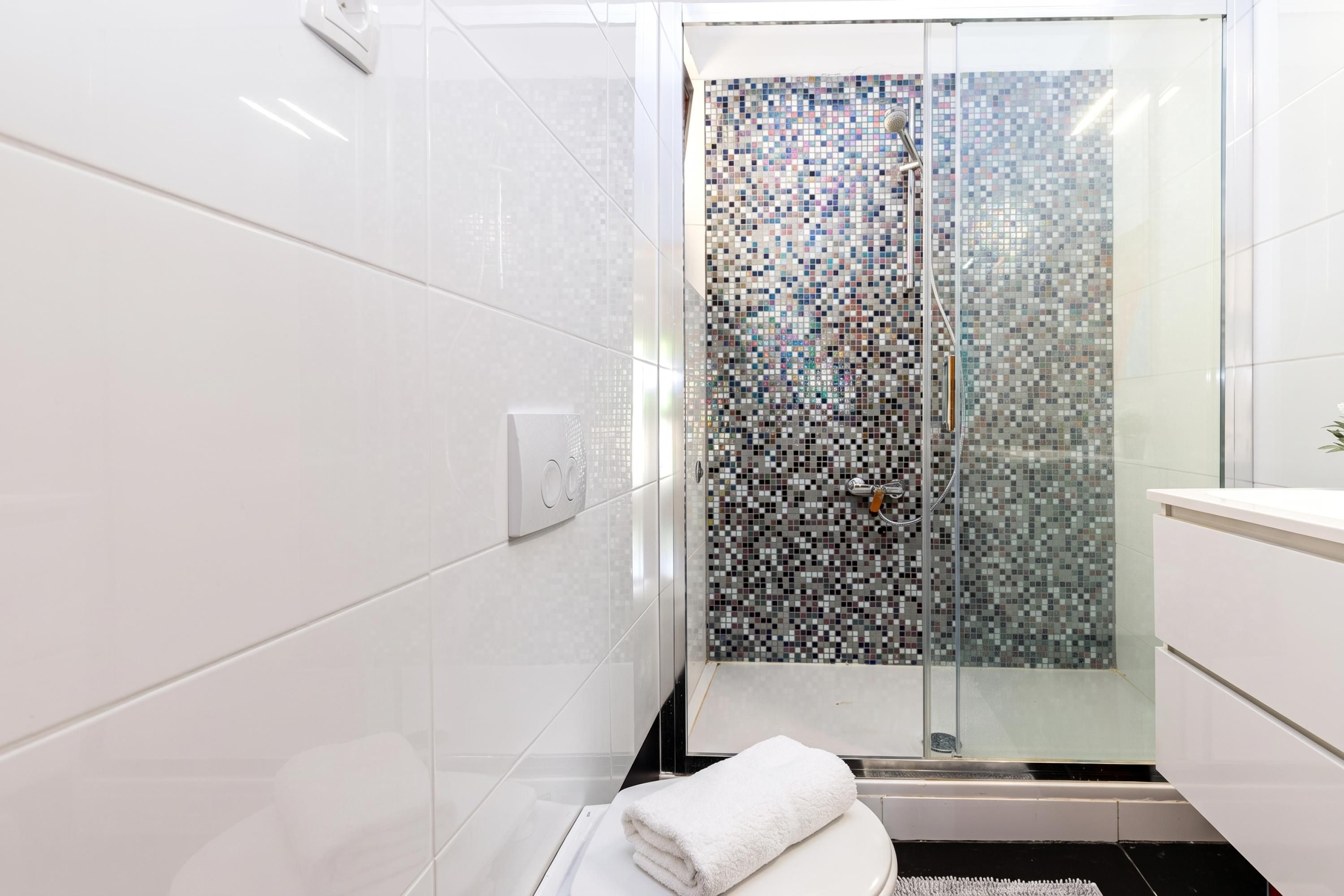 A modern bathroom with white tiles, a glass shower enclosure with mosaic tiles, and a white towel on the floor.