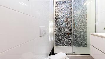 Un baño moderno con azulejos blancos, un box de ducha con mosaicos y una toalla blanca en el suelo.