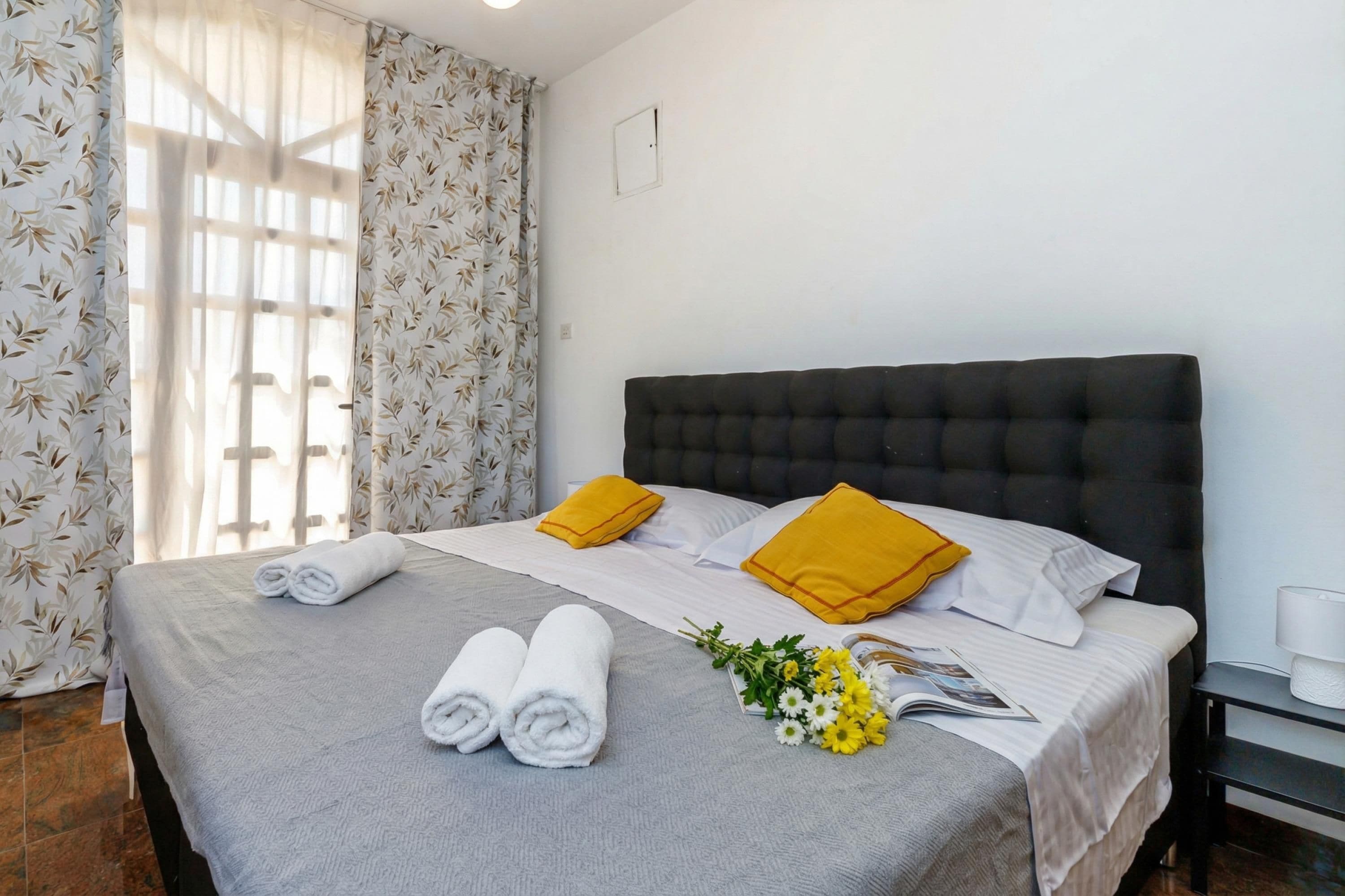 Una cama bien hecha con toallas enrolladas, dos almohadas, una revista y un pequeño ramo de flores.