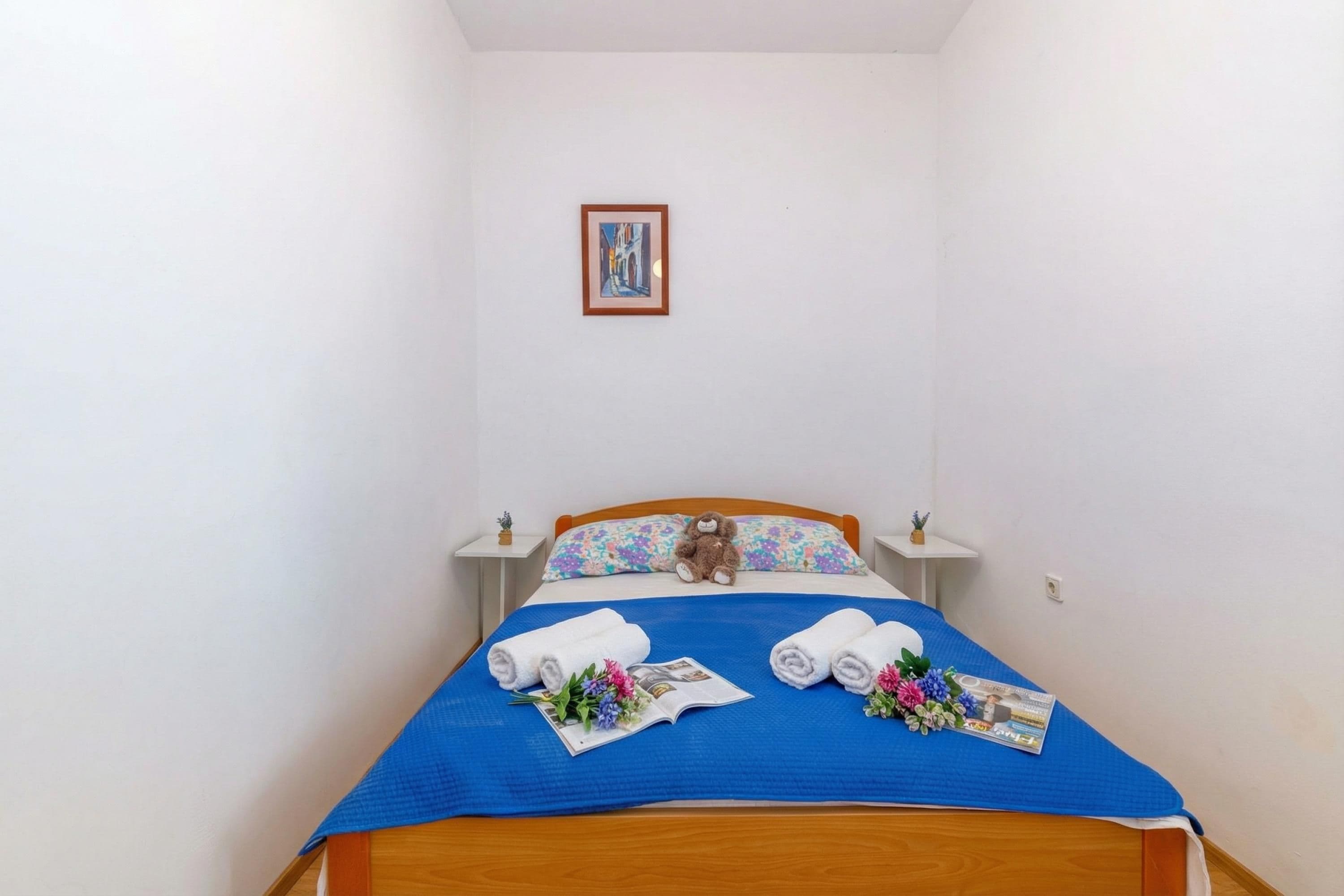 A neatly made bed with a blue bedspread, two pillows, a teddy bear, and rolled towels.