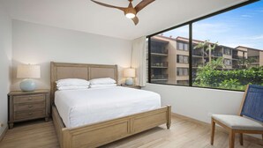 A hotel room with a wooden bed, two bedside lamps, a ceiling fan, a view of a building with balconies, and a chair. - Papakea Resort - Beautiful 1 BR Ocean View Suite - NO Resort Fee (Lahaina)