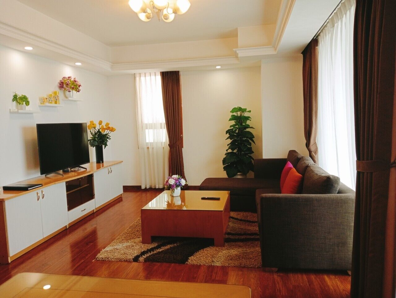 A modern living room with a flat-screen TV, a wooden coffee table, a grey sofa with red cushions, a large potted plant, and a white entertainment unit with shelves.