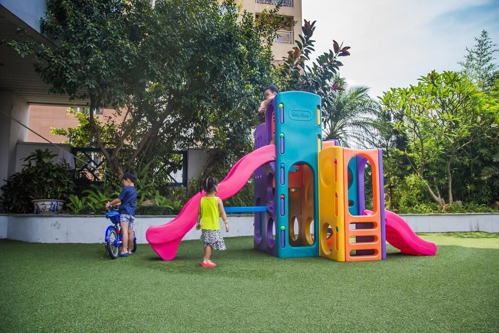 A playground with a slide and climbing structures, surrounded by greenery and a building in the background.