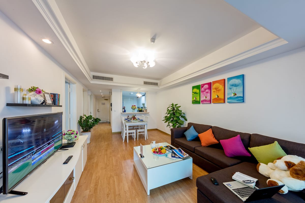 Ein modernes Wohnzimmer mit einem Flachbildfernseher, einem Couchtisch mit Früchten und Zeitschriften, einem braunen Sofa mit bunten Kissen und einem Essbereich im Hintergrund.