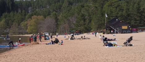 A beach with people engaging in various activities, a boathouse cafe, and a forested hillside in the background.