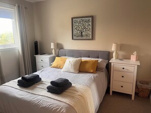 Un dormitorio ordenado con una cama, dos mesitas de noche, un buró, un cuadro en la pared y una ventana con cortinas.