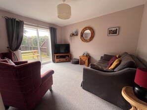A living room with a sofa, armchair, TV stand, and a view to the outside.