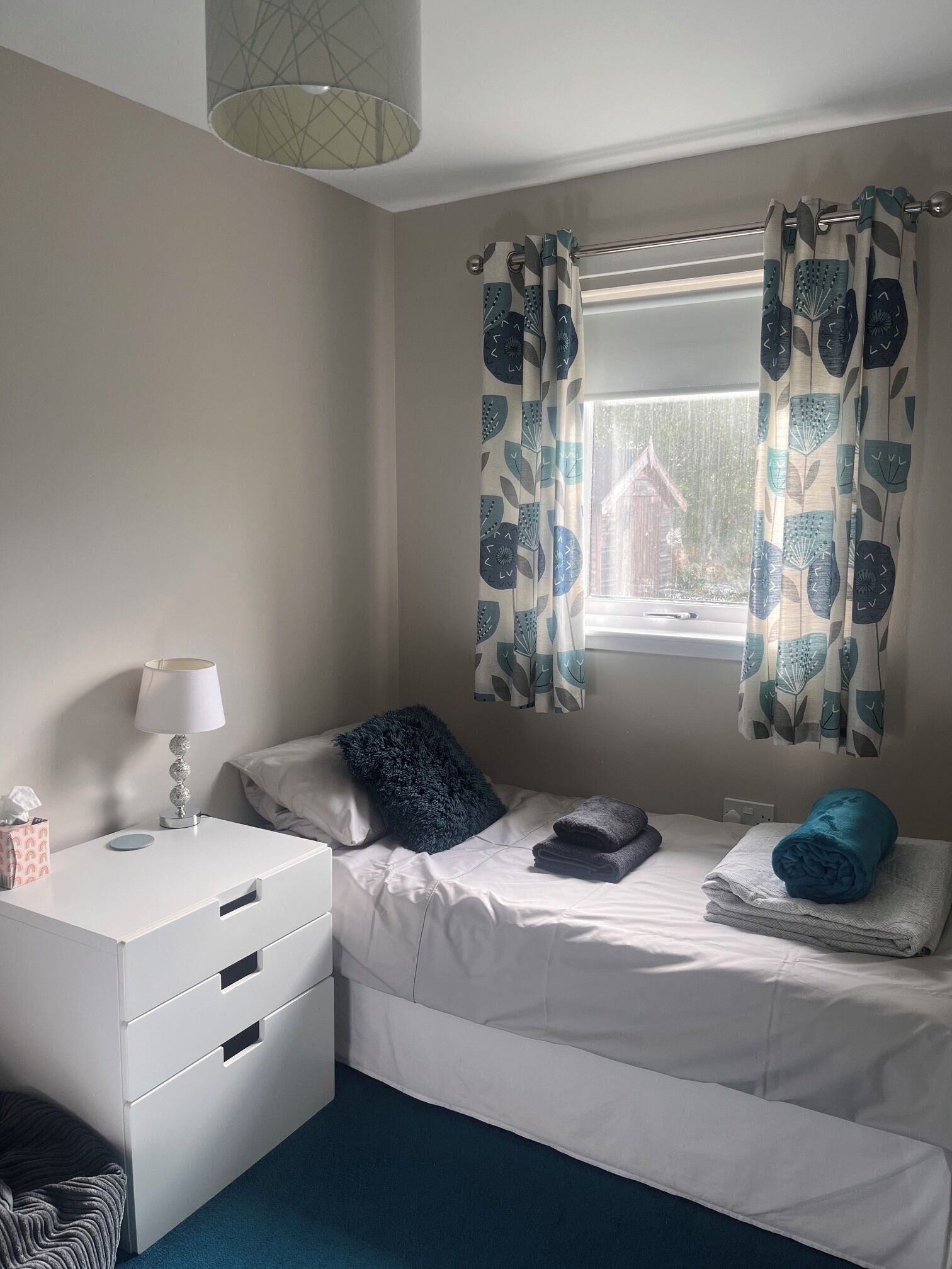 A bedroom with a bed, a bedside table with a lamp, a window with curtains, and a view of the outside.