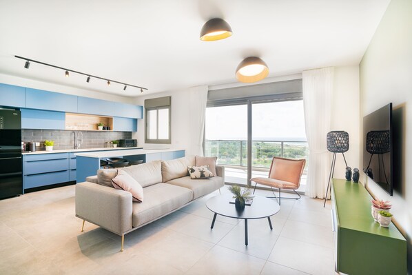 A modern living room with a sofa, coffee table, and a TV. There is a kitchen area with blue cabinets and a large window. - Sunny Haven Sea View Escape by Sea N Rent (Nahariya)