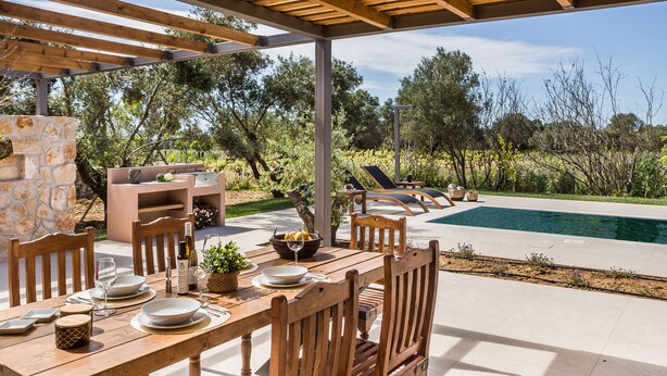 An outdoor dining area with a pergola, wooden chairs, a table set for four, and a pool in the background. - Moschato Estate Villas (Σβορωνάτα)