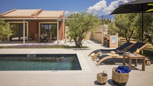 A poolside area with a clear pool, lounge chairs, a table, and a covered seating area. - Moschato Estate Villas (Σβορωνάτα)