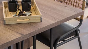 A wooden dining table with a tray holding two black mugs and a plate with cookies. A wicker chair is positioned at the table, and a brown sofa with cushions is in the background. - Moschato Estate Villas (Σβορωνάτα)