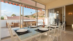 A dining area with a glass table and chairs, a bottle of wine, and a view of a mountain and palm trees.