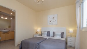 A bedroom with two beds, a nightstand with a lamp, a wall-mounted artwork, and a bathroom visible through an open door.
