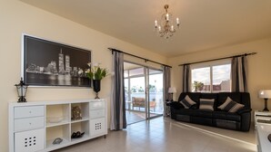 A spacious living room with a large flat-screen TV, a black leather sofa, a white console table, and a chandelier.