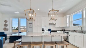 Una cocina moderna con armarios blancos, una isla grande con taburetes de bar, iluminación colgante y vistas al exterior.