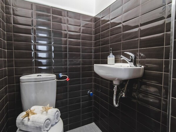 A bathroom with dark tiled walls, a white toilet, a sink with a faucet, and a towel on the floor.