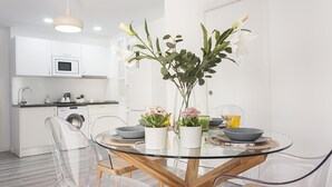 Een moderne keuken met witte kasten, een magnetron en een wasmachine. Er is een glazen eettafel met stoelen en een vaas met bloemen.