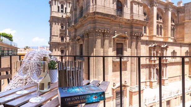 Een balkon met uitzicht op een historisch gebouw, een boek getiteld 'The Love Letter' van Lucinda Riley, en twee wijnglazen.