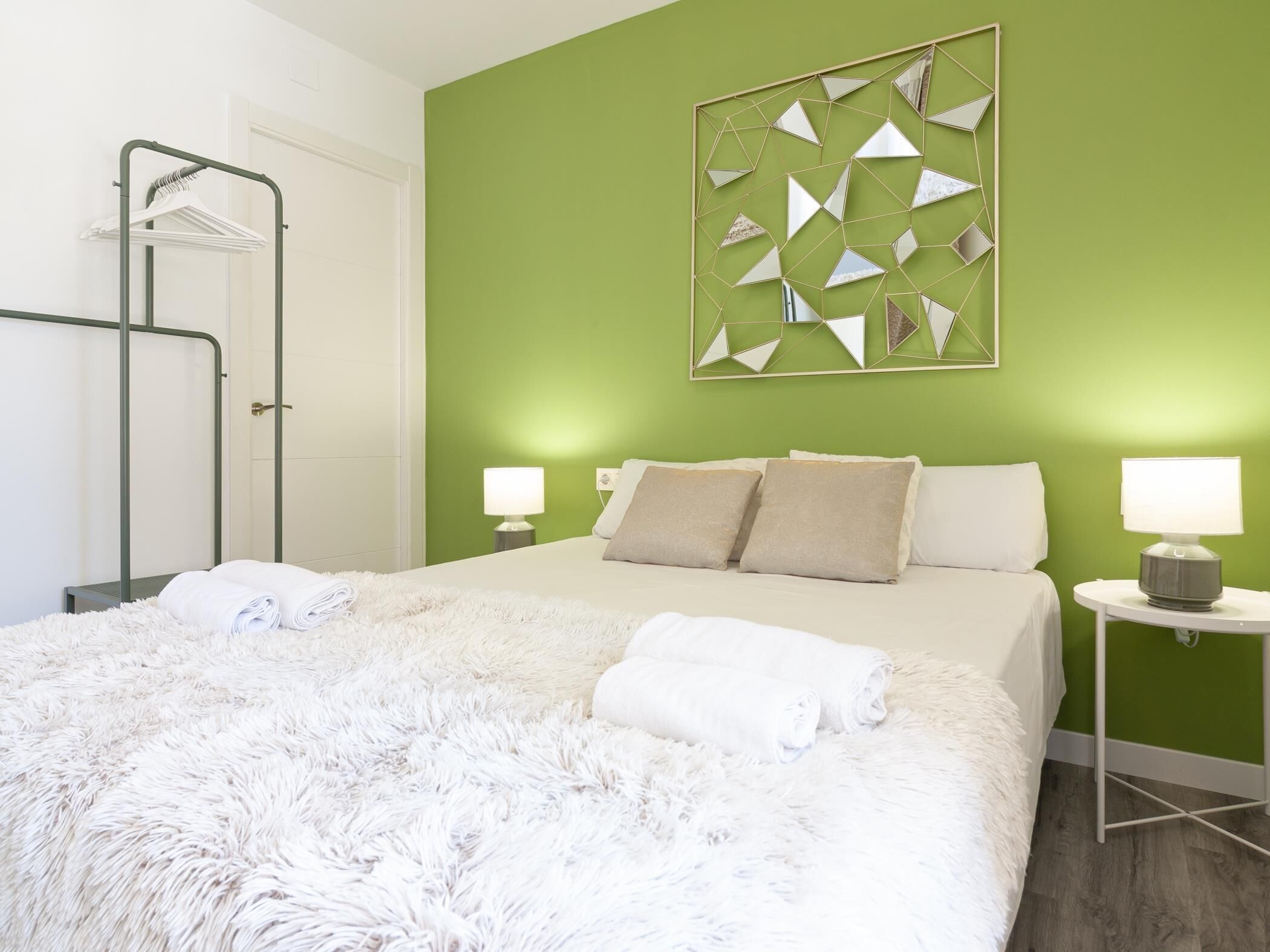 Una cama bien hecha con sábanas y ropa de cama blancas y mullidas, una pared verde con diseño de espejo geométrico y una mesita de noche con lámpara.